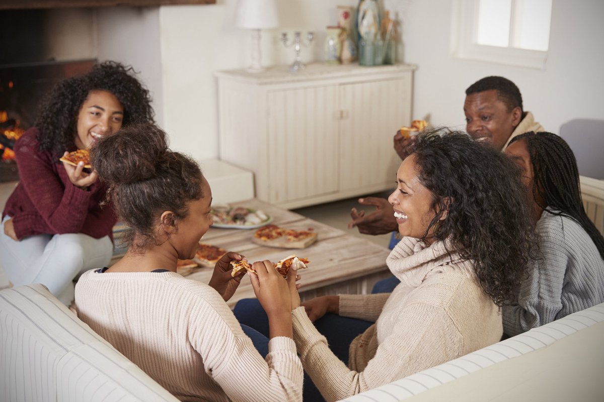 A family of five laughing together and eating pizza while sitting on the couch in their living room.