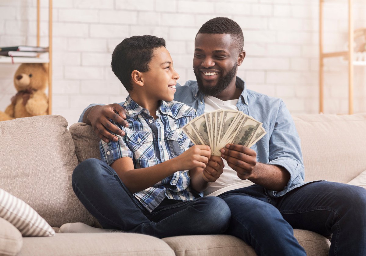 Father and son sit on the couch together holding cash.