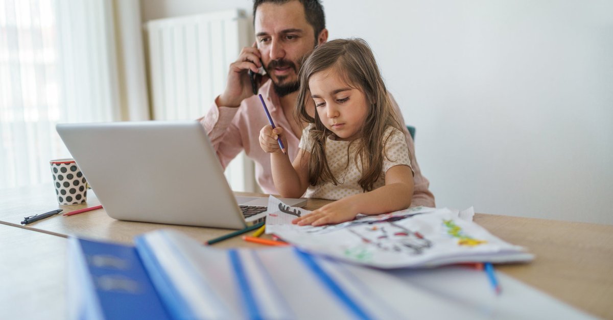 Father using laptop while on cell phone with young daughter coloring in his lap