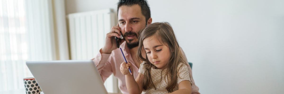 Father using laptop while on cell phone with young daughter coloring in his lap
