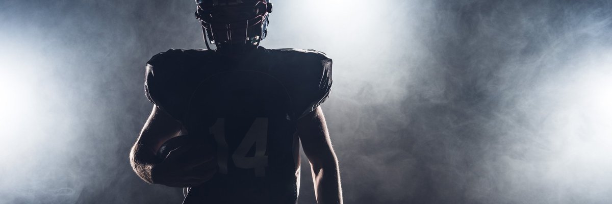 The silhouette of a football player in pads and a helmet holding a football.