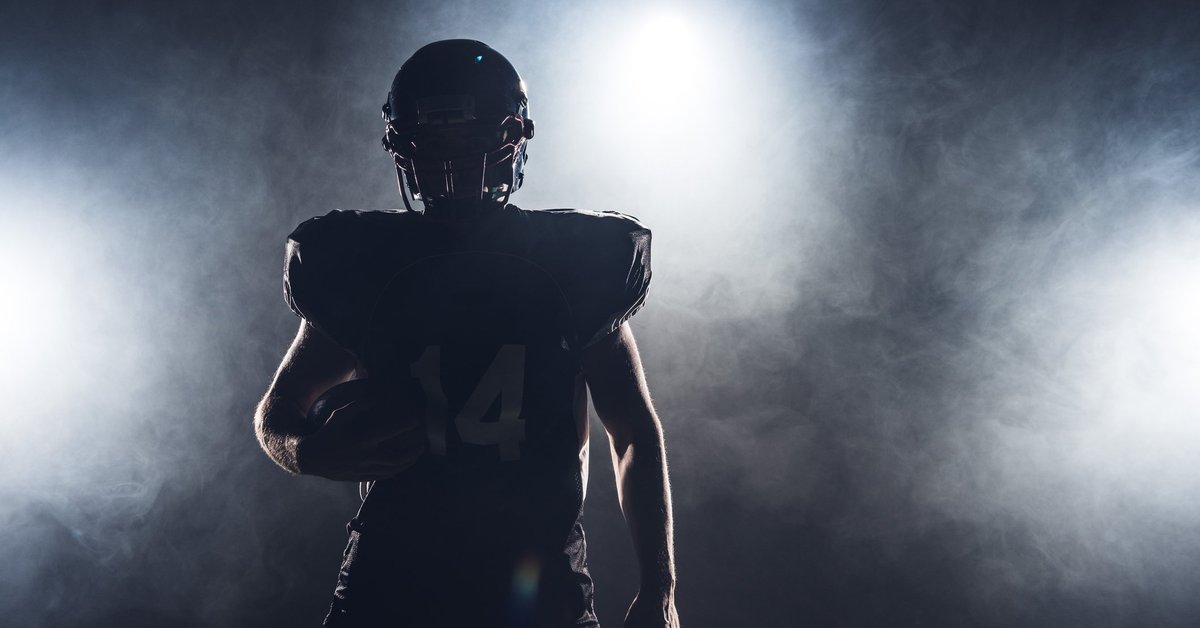 The silhouette of a football player in pads and a helmet holding a football.