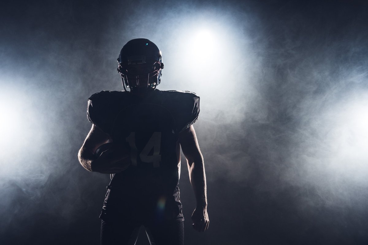 The silhouette of a football player in pads and a helmet holding a football.