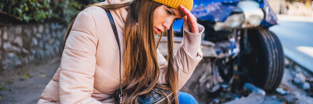 Frustrated young woman crouches next to wrecked car after a car accident.