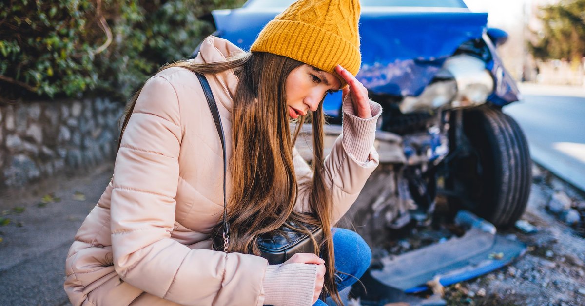 Frustrated young woman crouches next to wrecked car after a car accident.