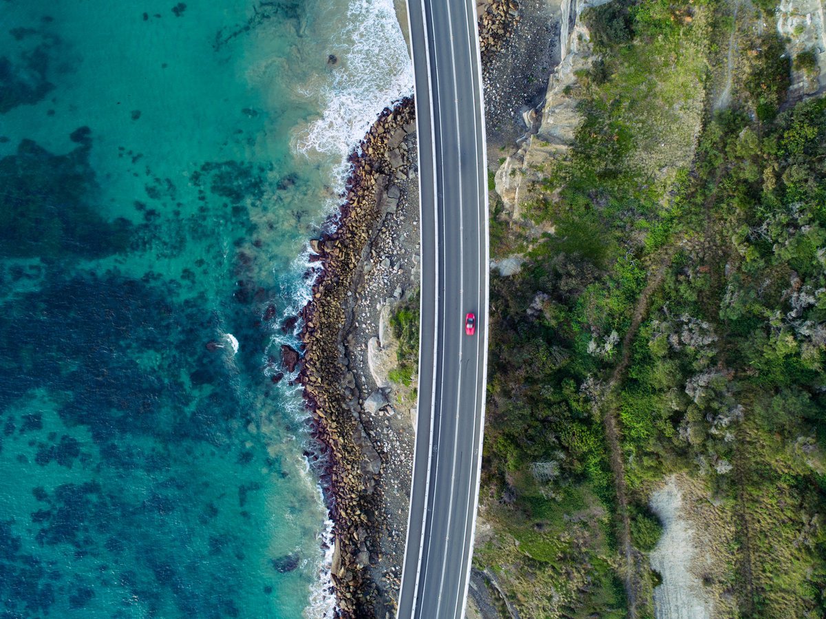Bird's eye view high above a shiny red car driving down a coastal highway.