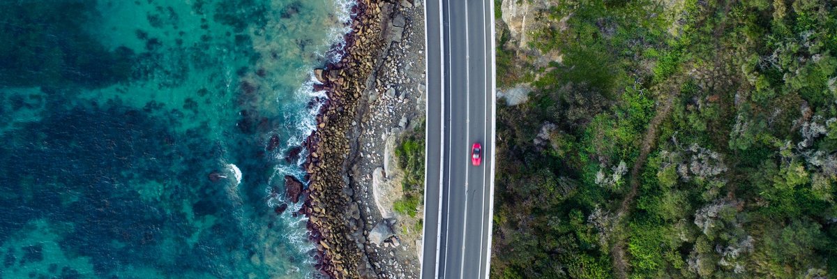 Bird's eye view high above a shiny red car driving down a coastal highway.