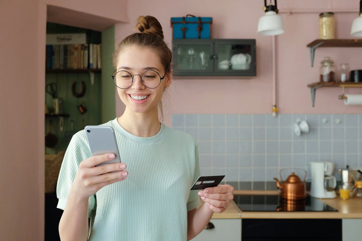 Smiling girl in kitchen with credit card and phone.
