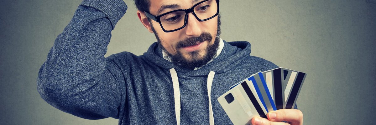 Bespectacled young man looking at the numerous credit cards in his hand with confused disbelief.