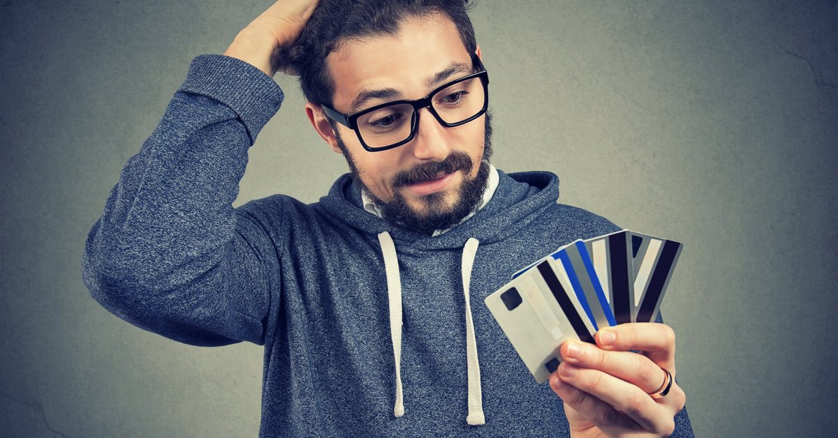 Bespectacled young man looking at the numerous credit cards in his hand with confused disbelief.