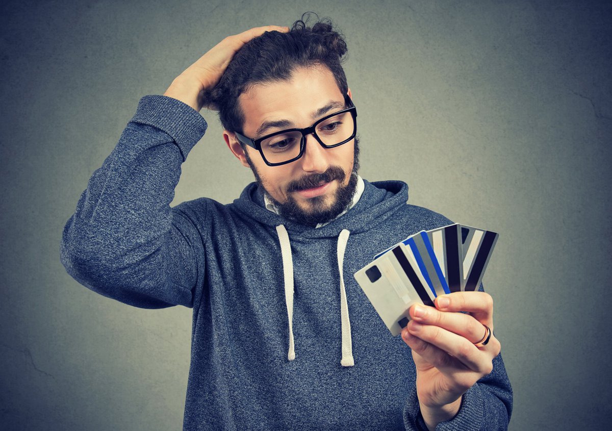 Bespectacled young man looking at the numerous credit cards in his hand with confused disbelief.