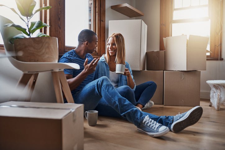 A happy couple looks sitting on the floor between moving boxes and drinking coffee.