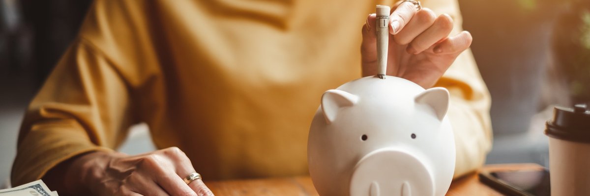 Person in a yellow sweater sitting behind desk and sliding a dollar bill into a piggy bank.