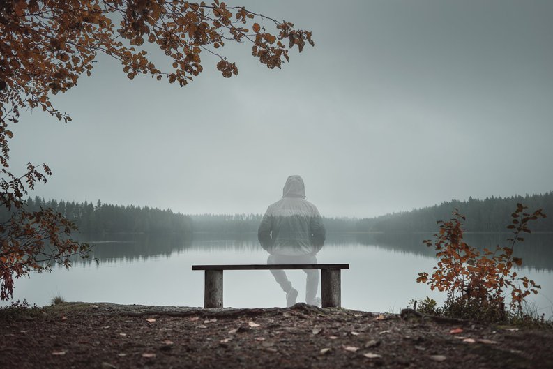 A man sitting on a bench looking out at a wintry lake and fading into physical transparency.