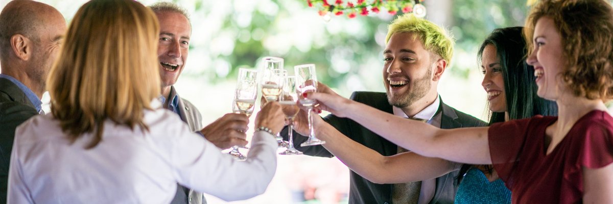 A group of wedding attendees in formal attire smile and toast their drinks.