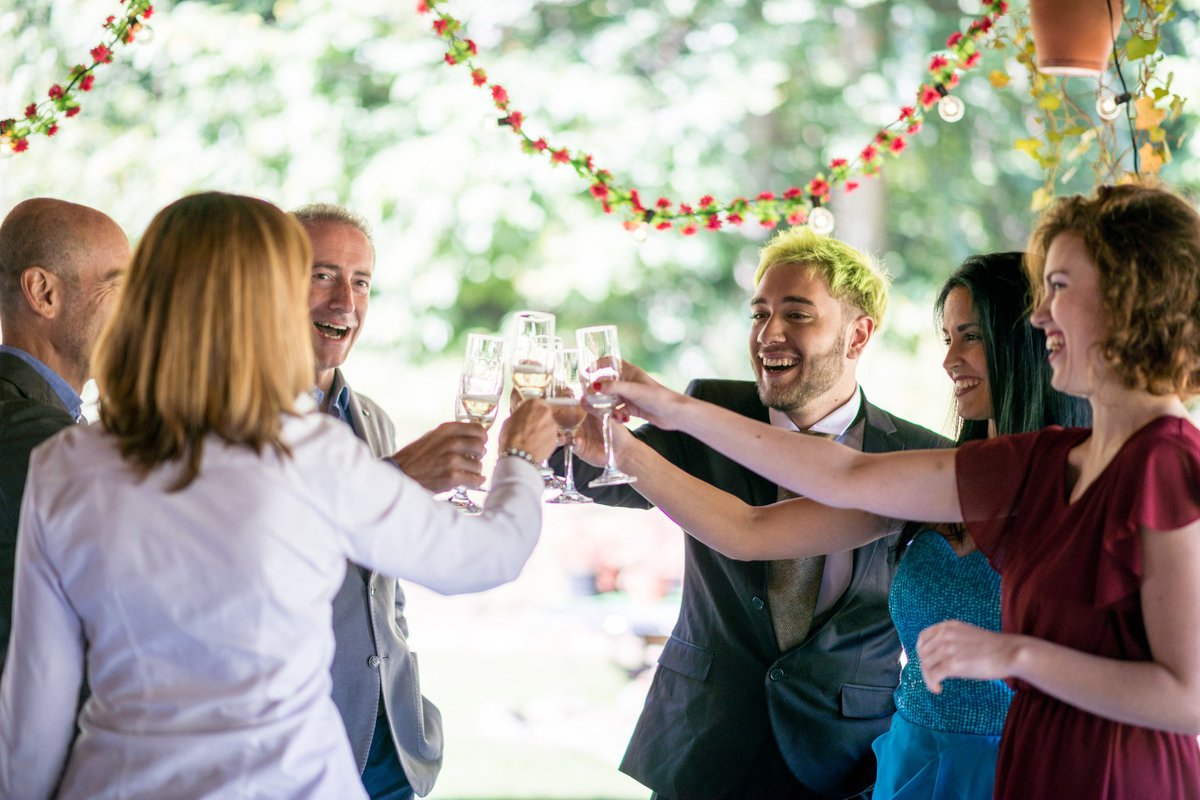 A group of wedding attendees in formal attire smile and toast their drinks.