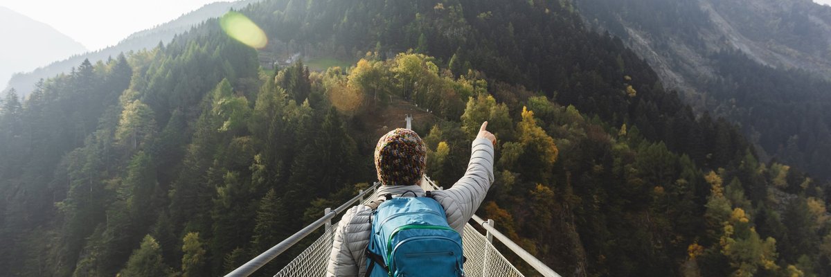 Man in puffer jacket and parka crossing narrow bridge over forest and pointing intrepidly at mountain in distance.