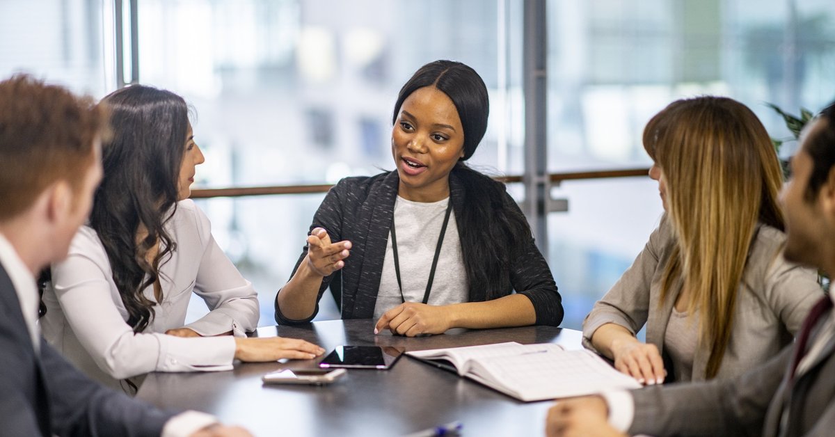 Young woman sitting at the head of a conference table leading a discussion with several co-workers.