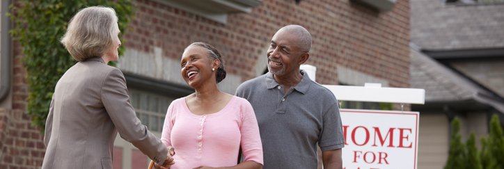 A realtor shakes hands with a senior couple in front of a For Sale sign and house.