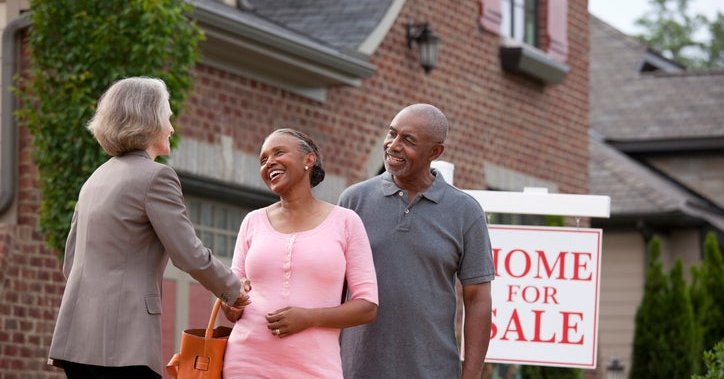 A realtor shakes hands with a senior couple in front of a For Sale sign and house.