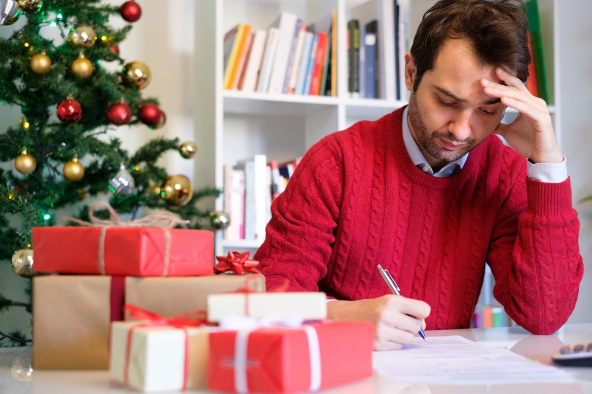 Guy in red sweater stressing over bills while surrounded by Christmas gifts and a Christmas tree.