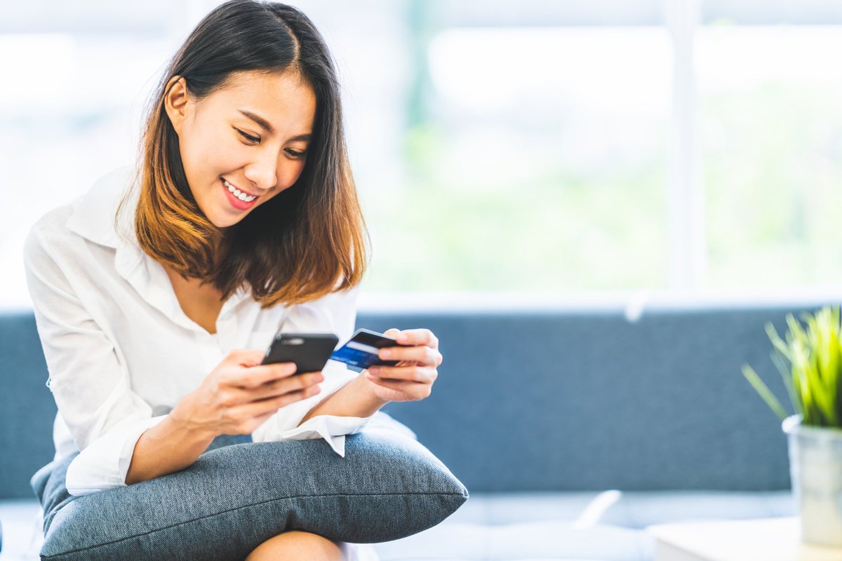Young woman smiles down at credit card in one hand and phone in the other.