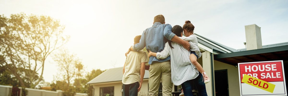 A family of four, arm in arm, look at a house with a "Sold" sign.