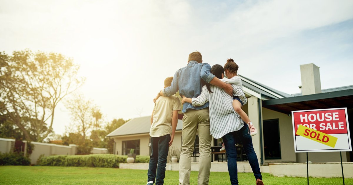 A family of four, arm in arm, look at a house with a "Sold" sign.