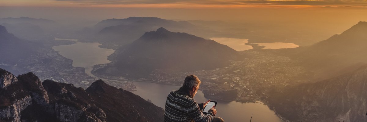 Middle-aged man looking at an iPad while sitting on rocks overlooking a sunset over mountains.