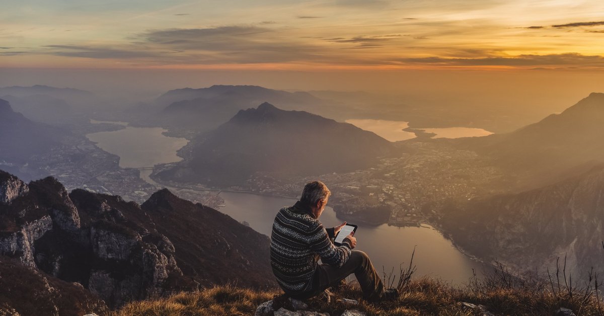Middle-aged man looking at an iPad while sitting on rocks overlooking a sunset over mountains.