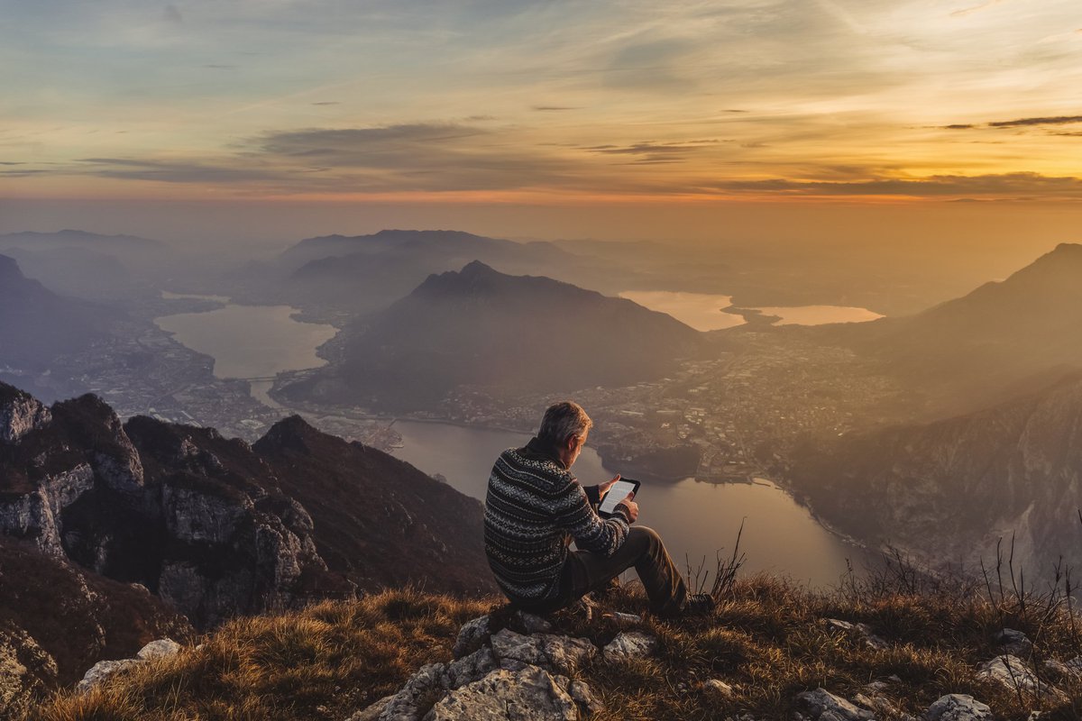 Middle-aged man looking at an iPad while sitting on rocks overlooking a sunset over mountains.