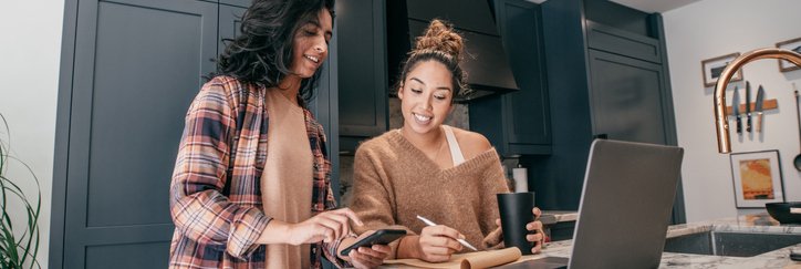 Two young adults wearing loungewear use their devices and a pad of paper to discuss finances.