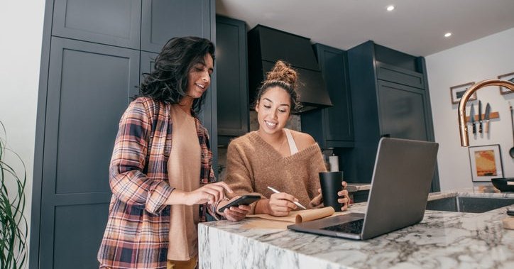 Two young adults wearing loungewear use their devices and a pad of paper to discuss finances.