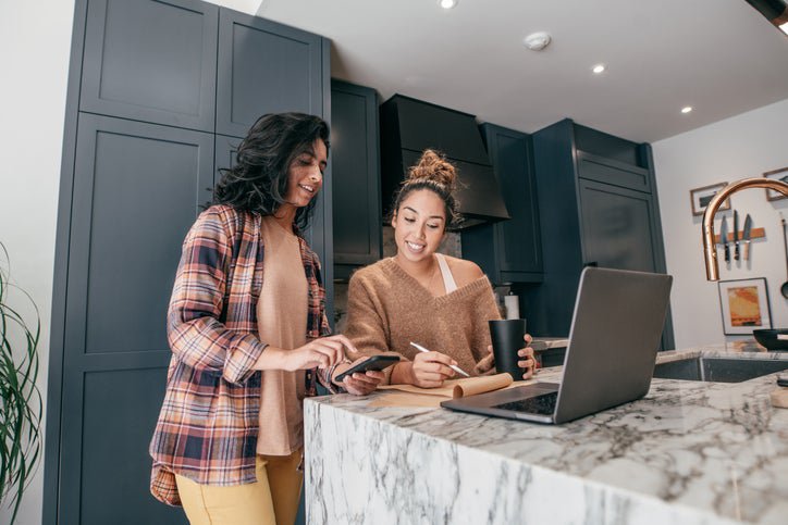 Two young adults wearing loungewear use their devices and a pad of paper to discuss finances.