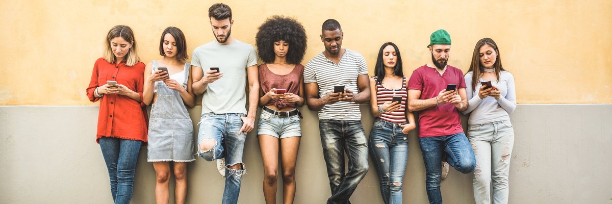 A bunch of young adults leaning against a wall on their phones.