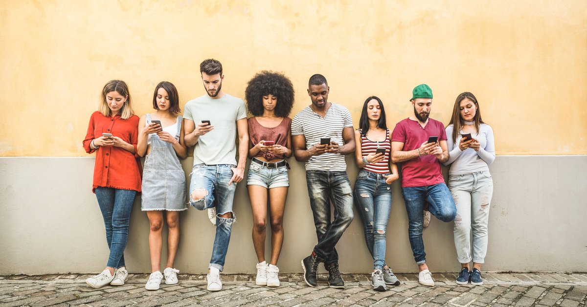 A bunch of young adults leaning against a wall on their phones.