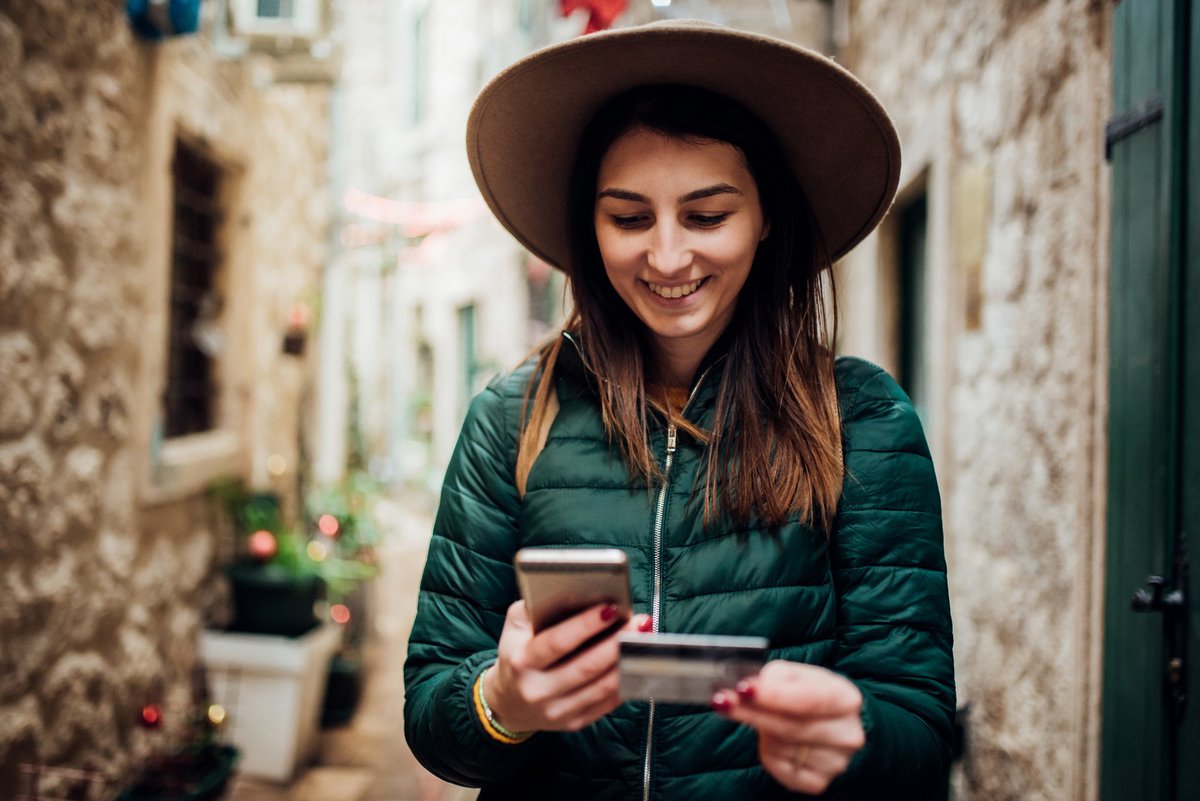 Young woman in hip wide-brimmed hat traveling through an alley in some ancient city grinning down at phone in one hand and credit card in the other.