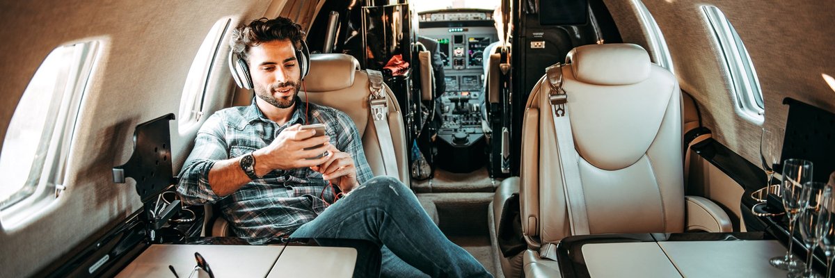 A person sitting in a private jet uses a smartphone and headphones while smiling.