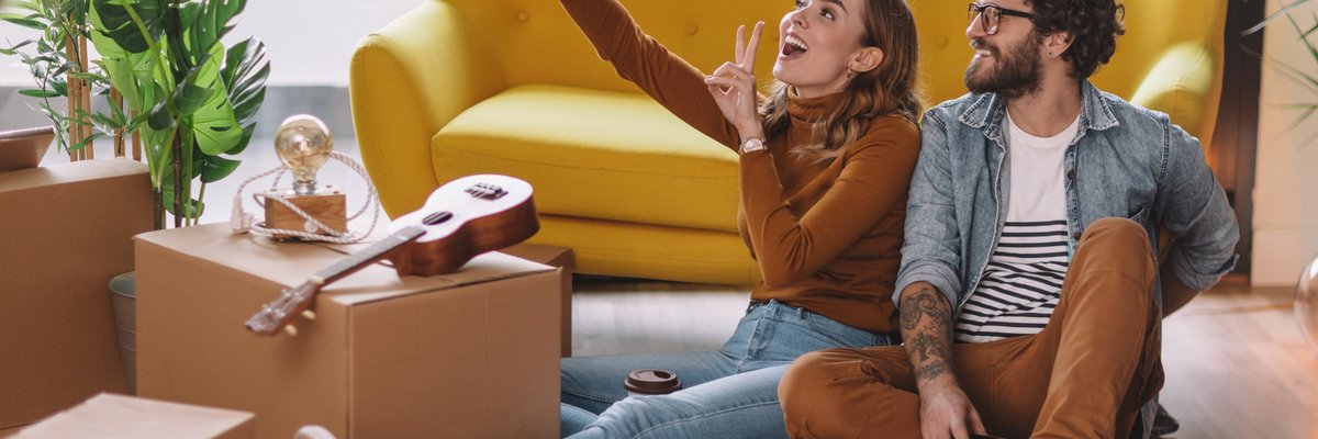 Two people take a selfie together while sitting on the floor next to moving boxes, a couch, and a ukulele.