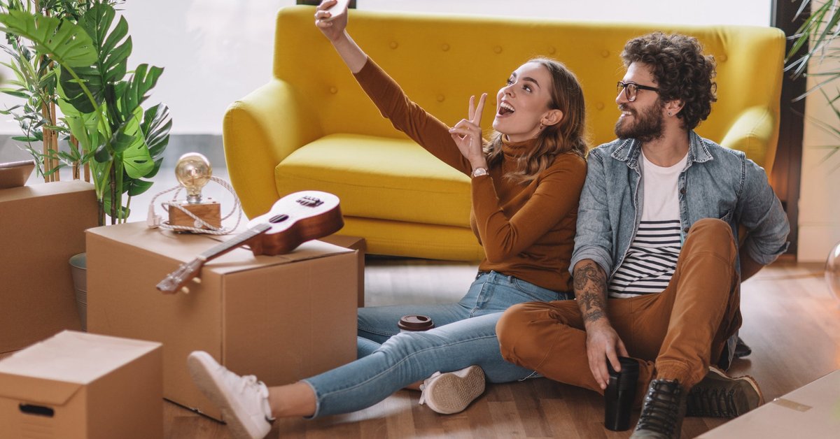 Two people take a selfie together while sitting on the floor next to moving boxes, a couch, and a ukulele.