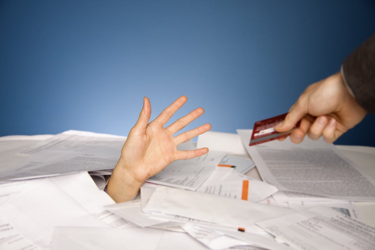 Hand extending credit card to other hand that's reaching out from beneath a pile of bills.