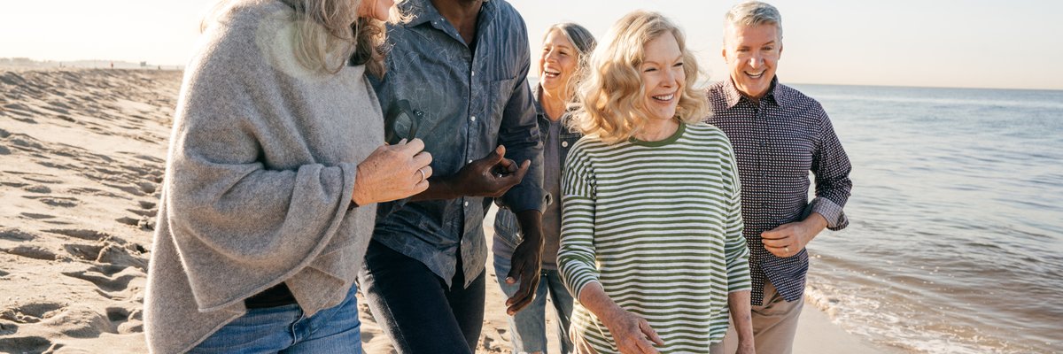 A group of people, some with gray hair, smile and converse as they walk along the beach.