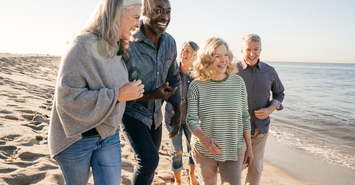 A group of people, some with gray hair, smile and converse as they walk along the beach.