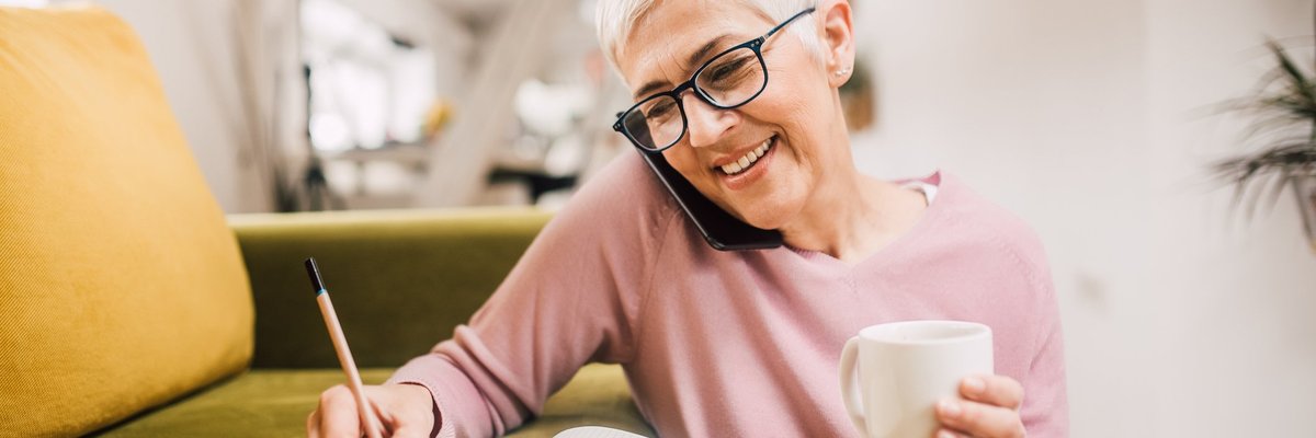 Older woman writing something down in notebook while on the phone.