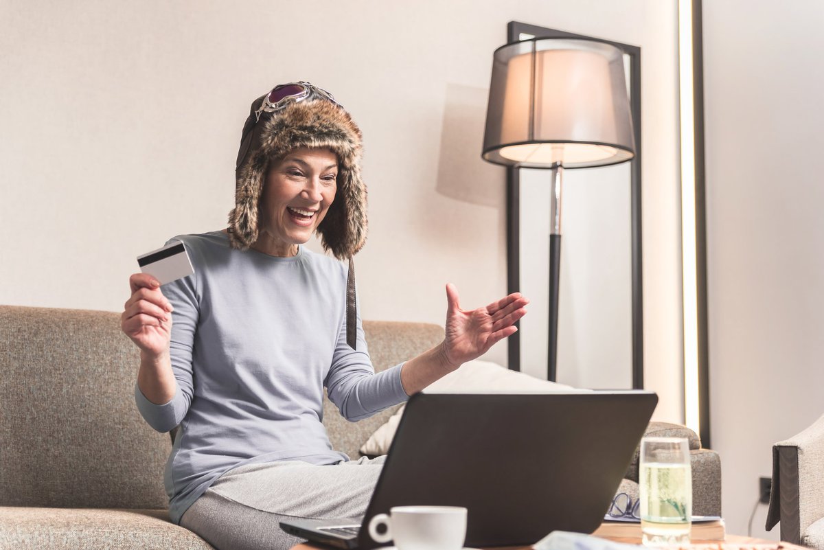 Woman in coonskin cap and goggles sitting on couch and beaming at laptop screen while holding credit card.