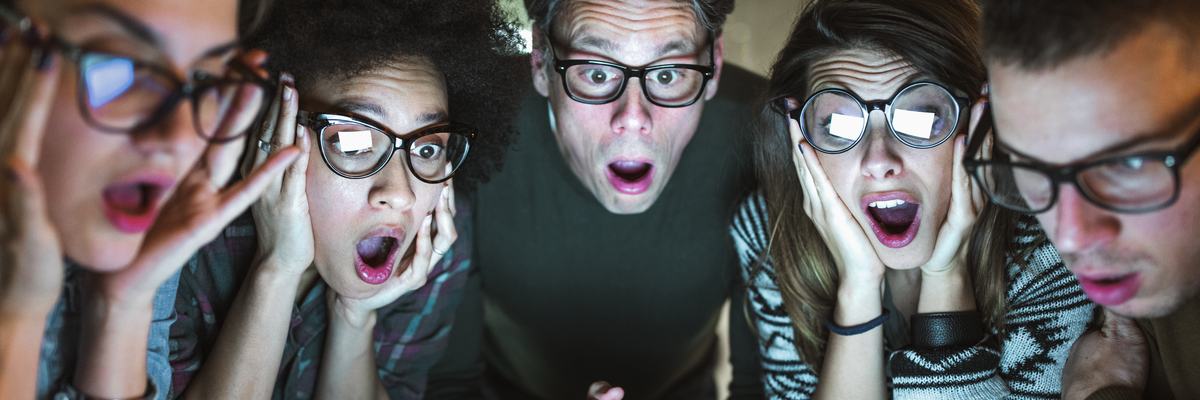 Group of adults in glasses crowd around laptop screen with hands to their faces in shock at what they see.
