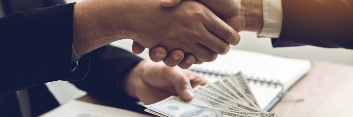 A pair of hands shaking hands and also transferring a wad of 100-dollar bills.