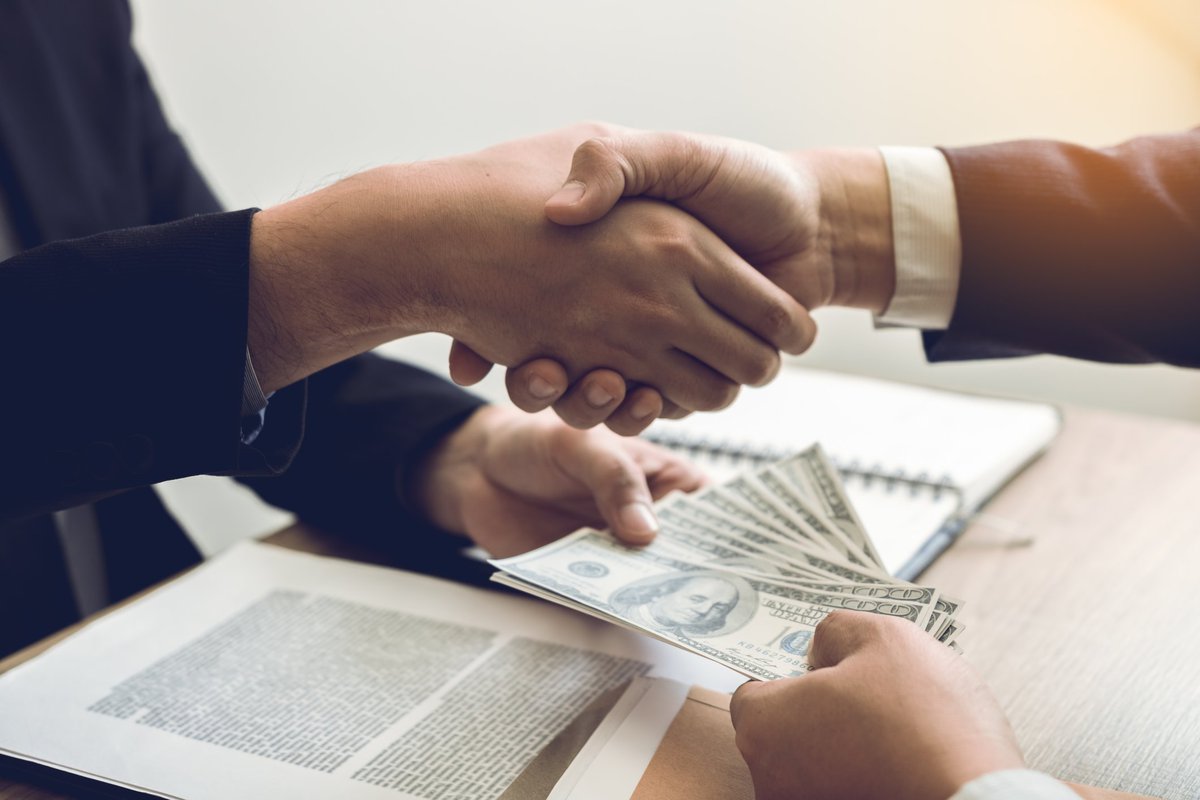 A pair of hands shaking hands and also transferring a wad of 100-dollar bills.