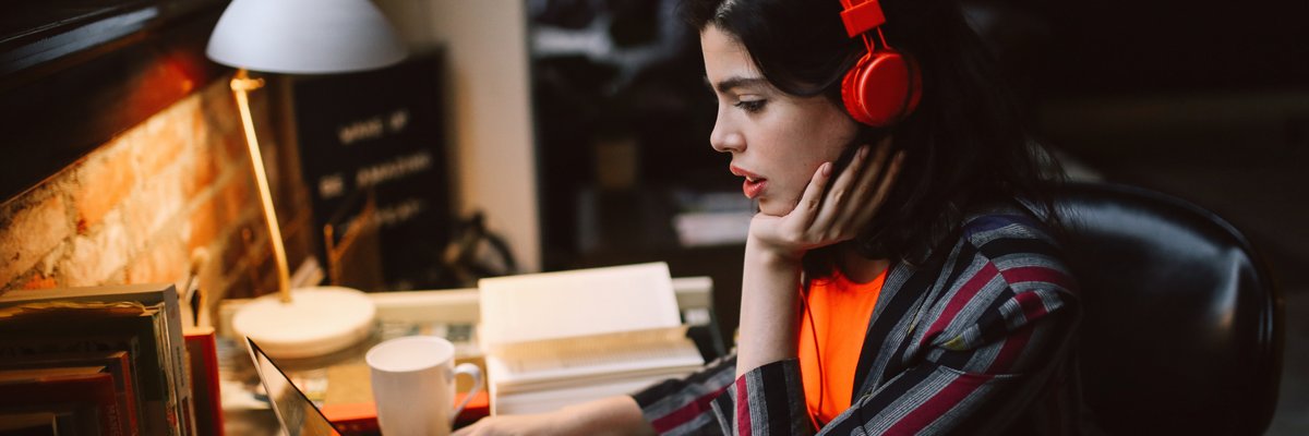 A person sits at a desk at home looking at a laptop screen and wearing headphones.