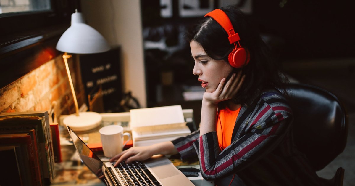 A person sits at a desk at home looking at a laptop screen and wearing headphones.